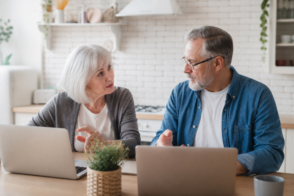 Older couple at a kitchen table, each using a laptop, discussing their email lists.