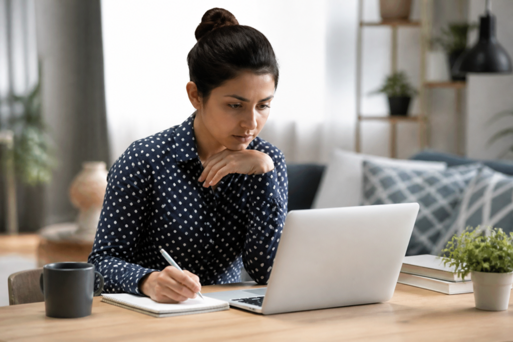 A woman sitting at a desk with a laptop, notepad, and coffee, diligently analyzing competitors in her chosen niche.